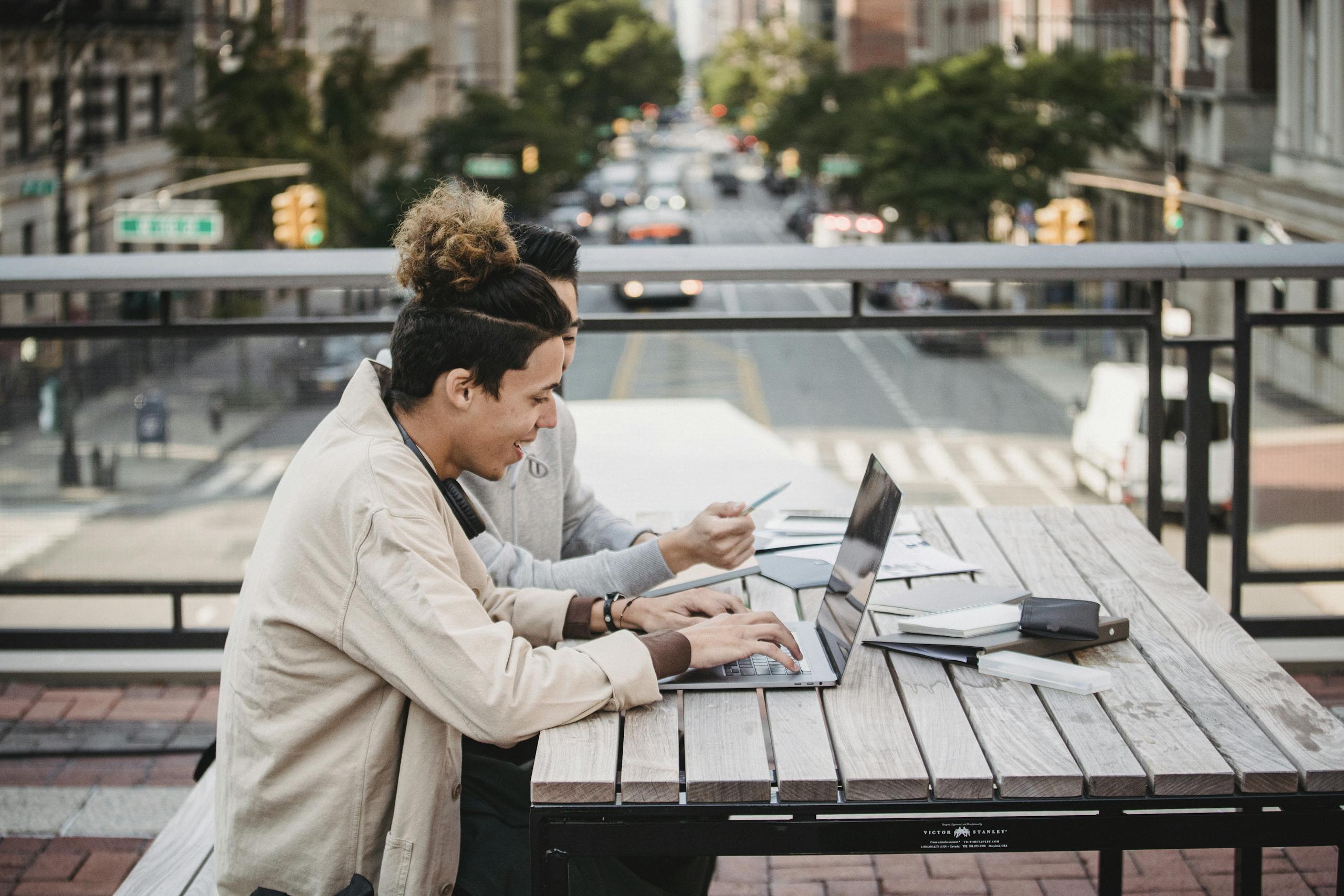 Side view of smiling young diverse male students in casual wear sitting at table with laptop and surfing internet while working on project together